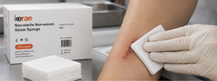 Gloved hand pressing a Kerae non-woven gauze sponge onto a sutured wound on a forearm